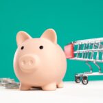Piggy bank with shopping cart and coins on a white and teal background, symbolizing saving and spending.
