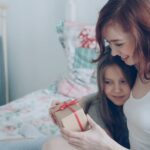 Mother and daughter joyfully exchange a wrapped gift on the bed.