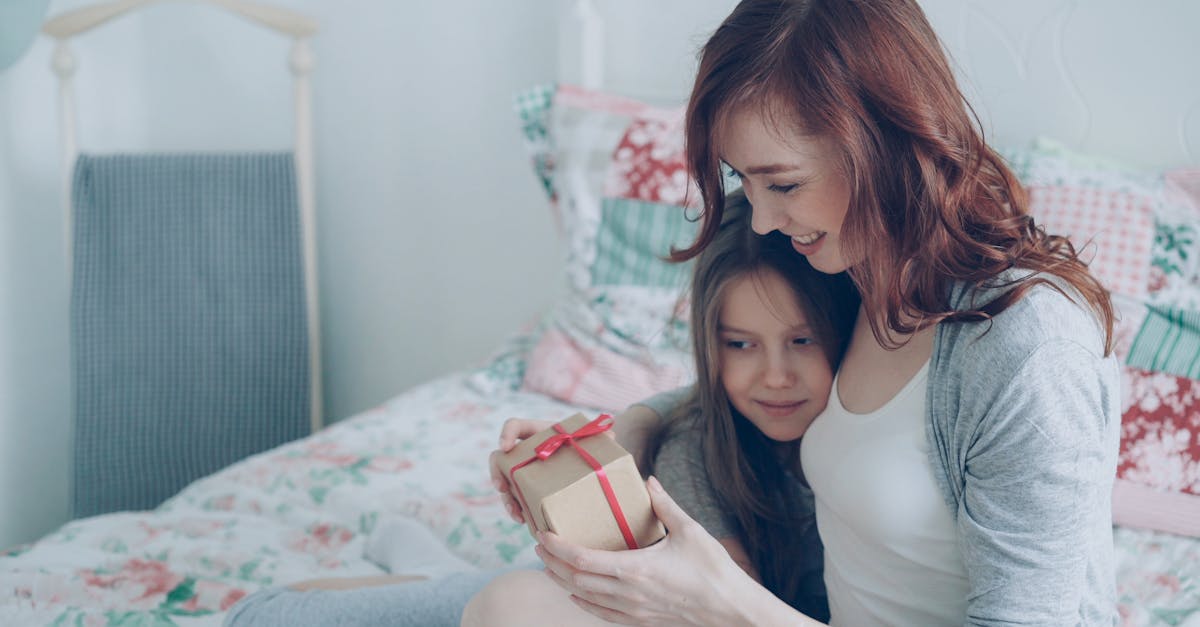 Mother and daughter joyfully exchange a wrapped gift on the bed.
