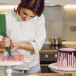 Focused young woman in uniform piping cream from pastry bag on middle of desserts after preparing delicious cake in modern cafeteria