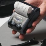 Close-up of a hand holding a digital printing device with a receipt, surrounded by grooming tools.