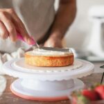 Close-up of person decorating a cake with frosting using a pink spatula on a cake stand.