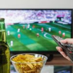 A person holding a remote while watching soccer on TV with beer and snacks on the table.