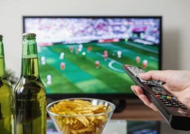 A person holding a remote while watching soccer on TV with beer and snacks on the table.