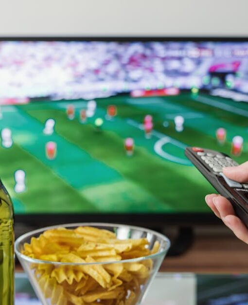 A person holding a remote while watching soccer on TV with beer and snacks on the table.