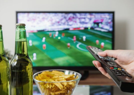 A person holding a remote while watching soccer on TV with beer and snacks on the table.