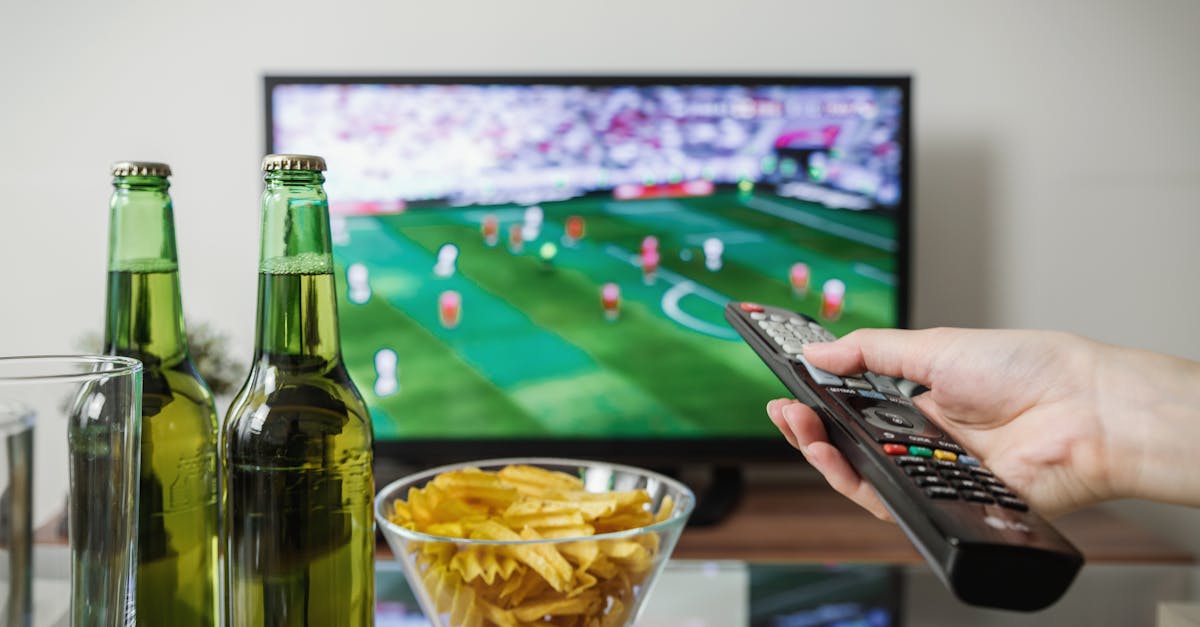 A person holding a remote while watching soccer on TV with beer and snacks on the table.