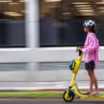 Woman in pink jacket rides a yellow electric scooter on a city street. Blurred motion effect.