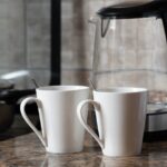 A warm indoor shot of two coffee mugs next to a kettle on a stylish kitchen counter.