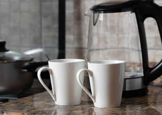 A warm indoor shot of two coffee mugs next to a kettle on a stylish kitchen counter.
