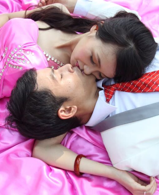 A couple in formal attire embraces on a pink blanket, enjoying a moment of affection outdoors.