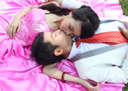 A couple in formal attire embraces on a pink blanket, enjoying a moment of affection outdoors.