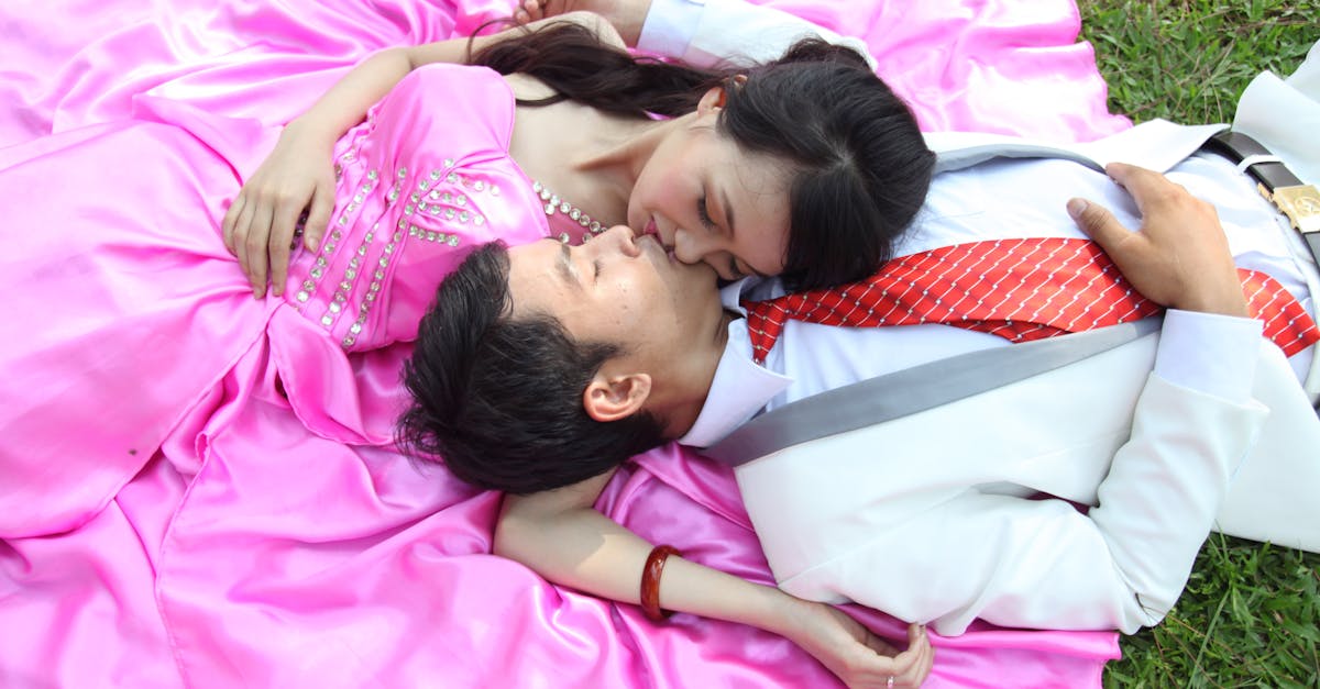 A couple in formal attire embraces on a pink blanket, enjoying a moment of affection outdoors.