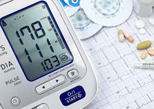 Close-up of a digital blood pressure monitor with ECG printout, pills, and medical tools.