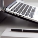 Side view of a laptop on a stand with a notebook and pen on a white desk.