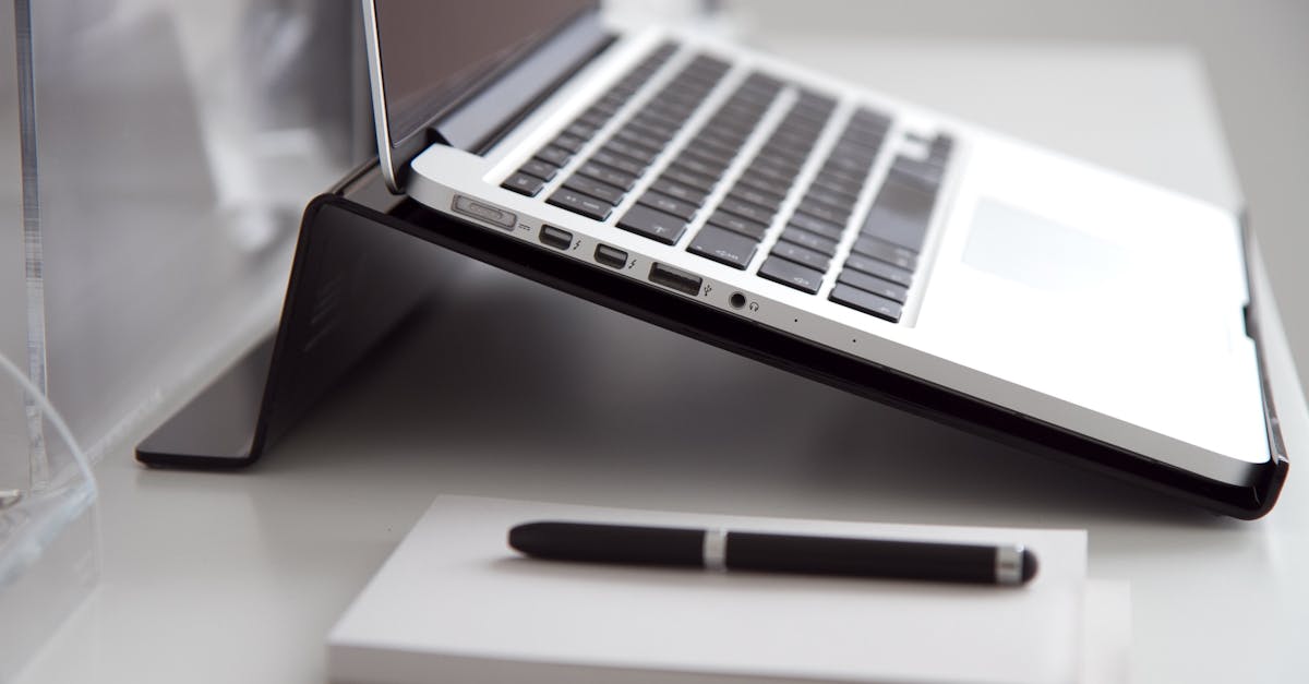 Side view of a laptop on a stand with a notebook and pen on a white desk.