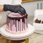 Unrecognizable female in apron and latex gloves garnishing delicious cake with sweet details while standing near table in modern restaurant