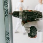 A construction worker uses a drill on drywall with blurred workers in the background.