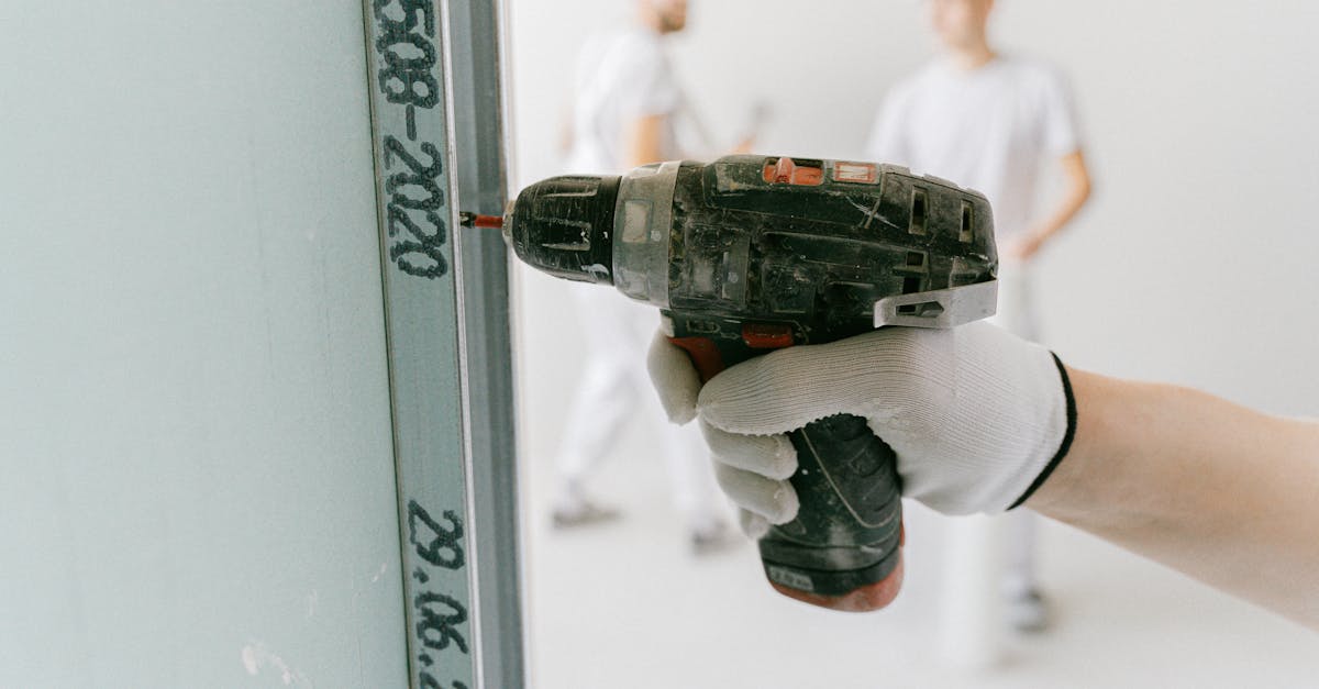 A construction worker uses a drill on drywall with blurred workers in the background.