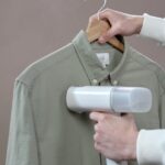 Hands steaming a green shirt with a portable steamer against a grey background.