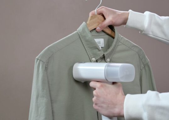 Hands steaming a green shirt with a portable steamer against a grey background.