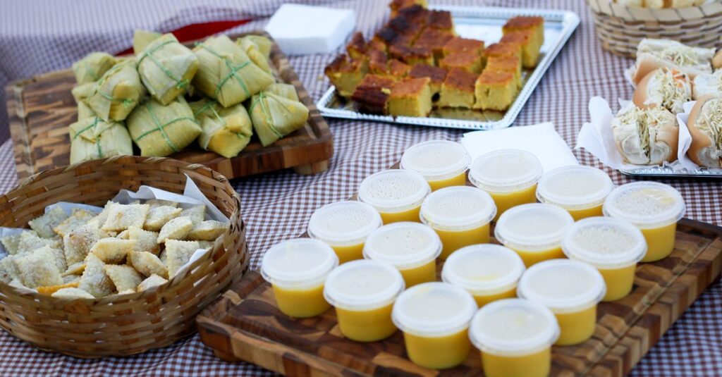 Tradições Juninas: Os Sabores que Mantêm Viva a Cultura das Festas de São João An assortment of traditional dishes including corn cake, tamales, and dessert cups on a checkered table.