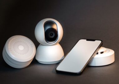 A sleek smartphone with modern smart home devices on a minimalist gray background.