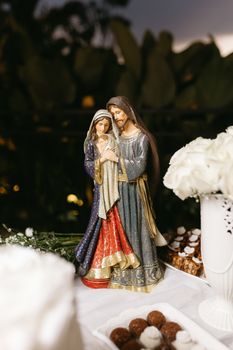 Sagrada Família de Nazaré: Um Símbolo de Amor, Fé e União para os Lares Cristãos A detailed nativity figurine of Mary, Joseph, and baby Jesus surrounded by white flowers and sweets.