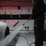 African American man cleaning a white car with a pressure hose in an indoor garage setting.