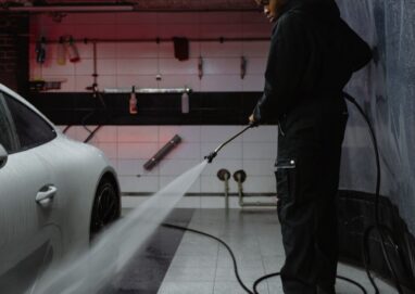 African American man cleaning a white car with a pressure hose in an indoor garage setting.