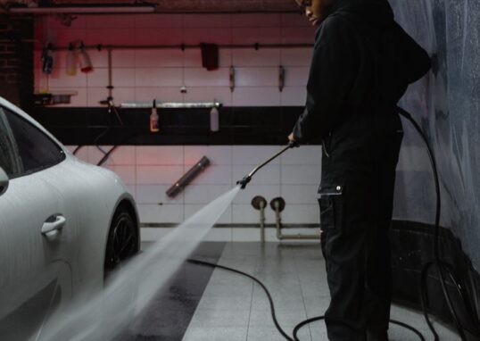 African American man cleaning a white car with a pressure hose in an indoor garage setting.