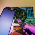 Hands holding a smartphone and remote control in a high-tech home setup with TV and other electronic devices.