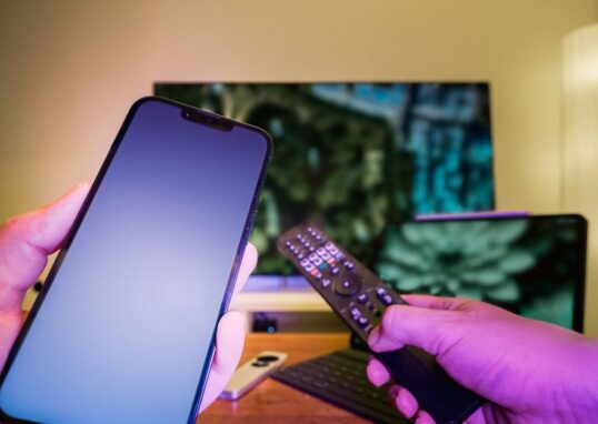 Hands holding a smartphone and remote control in a high-tech home setup with TV and other electronic devices.